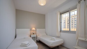 A small, clean room with two single beds, a bedside table with a lamp, a window with curtains, and a white chair.