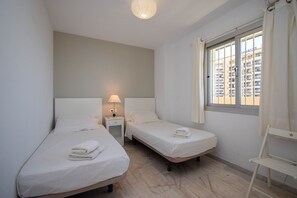 Ein kleines, sauberes Zimmer mit zwei Einzelbetten, einem Nachttisch mit Lampe, einem Fenster mit Vorhängen und einem weißen Stuhl.