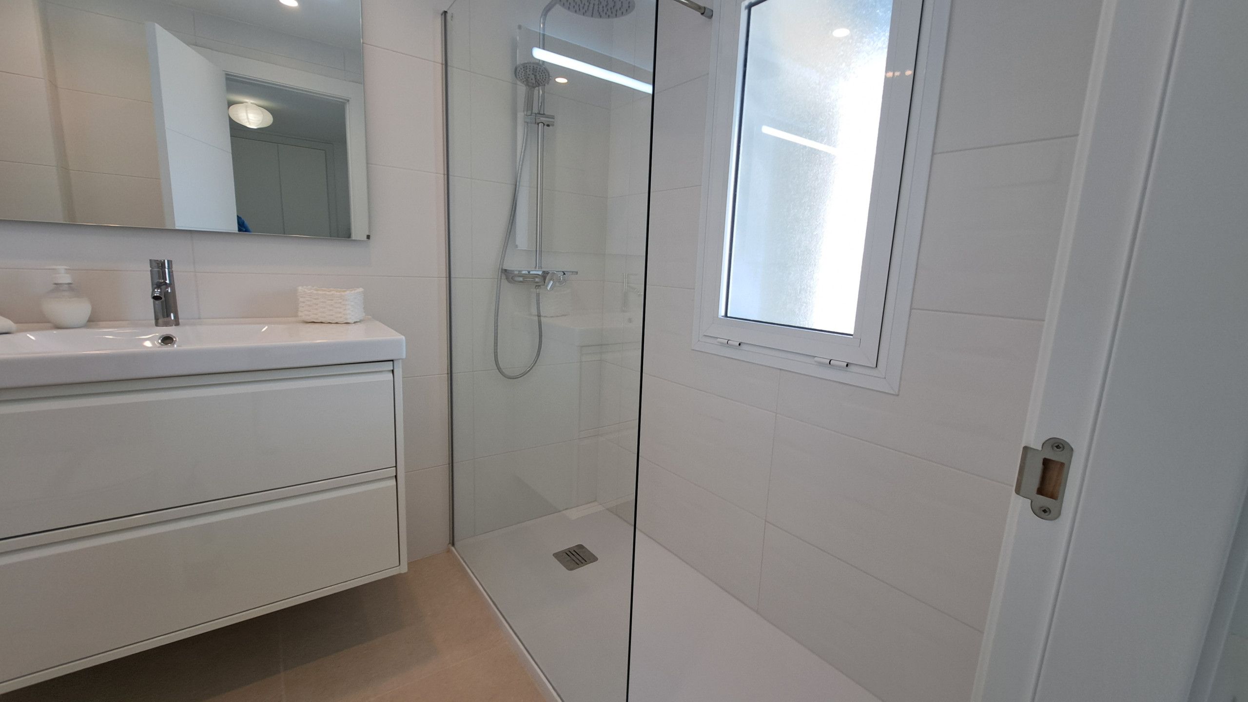 A modern bathroom with a glass-enclosed shower, a white vanity with a mirror, and a window.