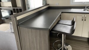A modern kitchen with a built-in microwave, a bar stool, and a window with curtains. - Rambling Rose caravan with sea view on Rockley Park (Poole)