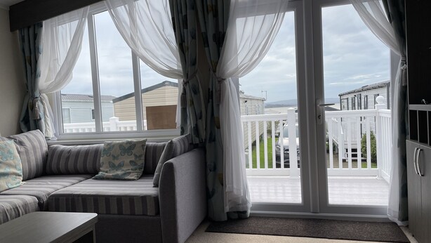 A modern living room with a gray sectional sofa, a small table, and sheer curtains. - Rambling Rose caravan with sea view on Rockley Park (Poole)