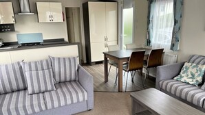 A modern living room with a sofa, a coffee table, and a dining area with a table and chairs. - Rambling Rose caravan with sea view on Rockley Park (Poole)