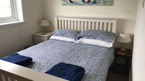 Una cama bien hecha con una colcha estampada, dos almohadas y dos toallas dobladas. En la pared sobre la cama, hay un cuadro con casas coloridas.