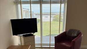 Un salón con televisor sobre un mueble bajo, una mesa de centro de madera, un sillón rojo y un ventanal con persianas.