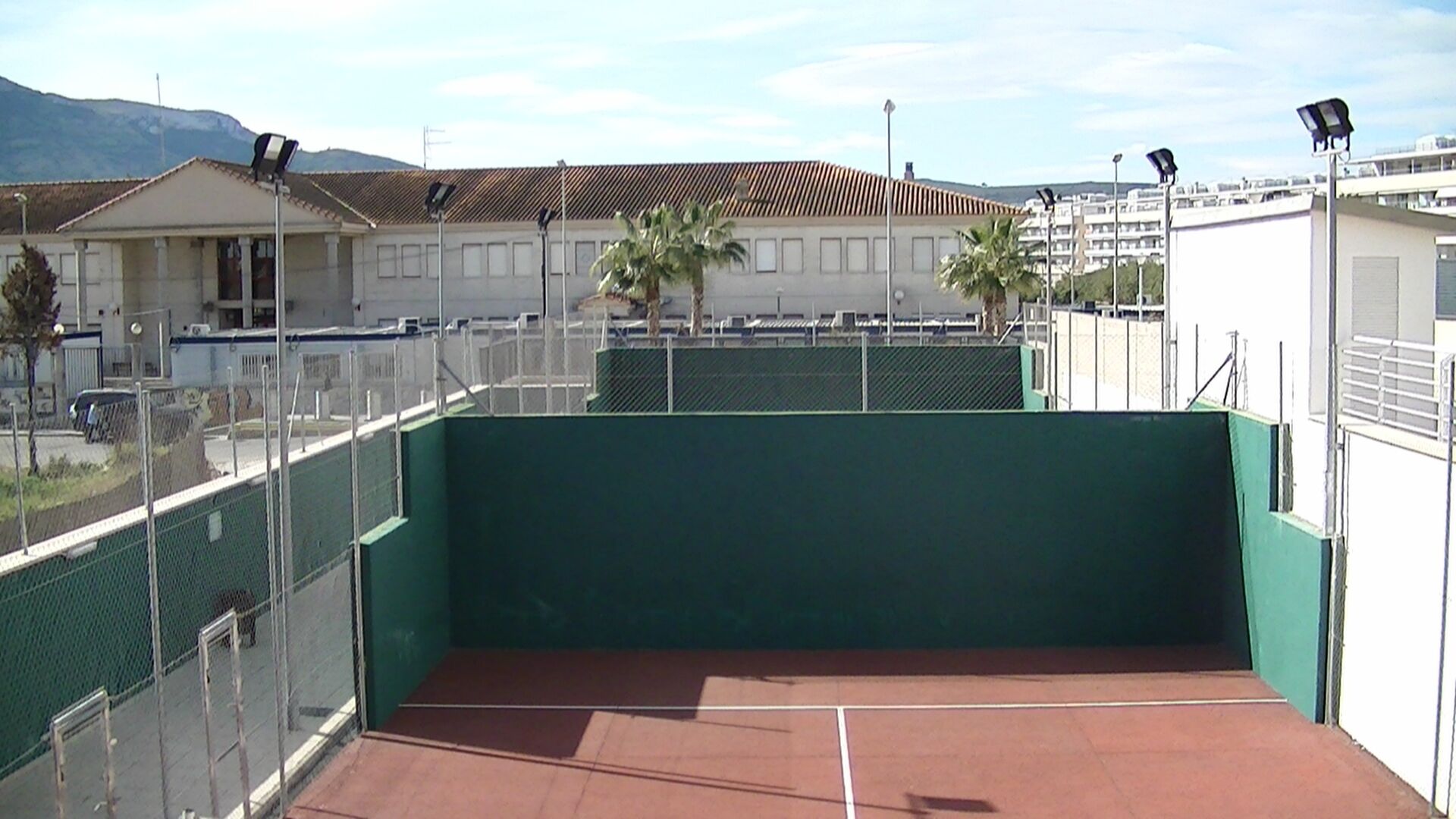 Una cancha de tenis con valla verde, un edificio al fondo y cielo despejado.
