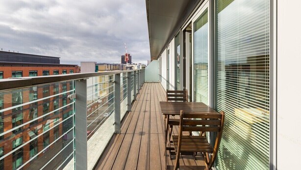A balcony with a wooden table and chairs, large windows with blinds, and a view of buildings in the background. - Skyline Birmingham City Centre Penthouse (Birmingham)