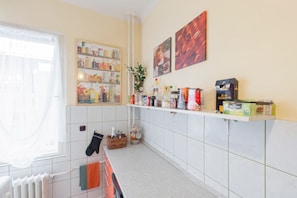 Una cocina con azulejos blancos, una ventana con cortinas traslúcidas, una estantería con diversos utensilios de cocina y un radiador.