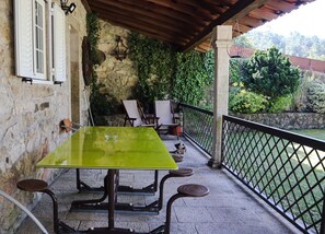 A covered outdoor seating area with a table and chairs, a stone wall, and a view of greenery.
