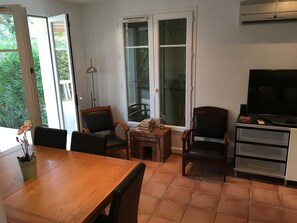 Et spiseområde med et træbord og stole, et lille sofabord med bøger og en lampe, og et fjernsyn på et stativ.