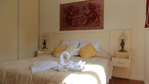 Een net opgemaakt bed met witte lakens en een witte handdoek, twee nachtkastjes met lampen, en een groot wandtapijt.
