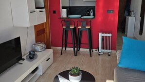 Ein modernes Wohnzimmer mit Fernseher, Unterhaltungseinheit und Couchtisch mit Pflanze. Eine rote Akzentwand, eine Küchenzeile mit Esstisch und Stühlen sowie ein graues Sofa mit blauem Kissen.