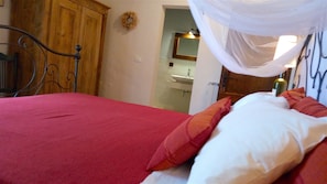 Une chambre à coucher avec un lit, une armoire en bois, une salle de bain visible à travers une porte ouverte et un baldaquin blanc au-dessus du lit.