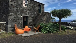 A stone building with a staircase leading to the entrance, outdoor seating with orange cushions, and a large tree in the foreground.