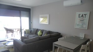 A modern living room with a grey sofa, a dining table with chairs, and a large window with blinds. - Holiday Houses - Los Altos 92 (Torrevieja)