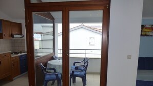 A kitchen with wooden cabinets and a stove, a balcony with a table and chairs, and a view of a building with a red roof. - APARTMENT GROZIĆ **** PORAT - KRK  (MALINSKA)