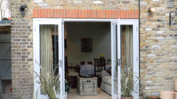 A brick house with a sliding glass door leading to a living room. There are two potted plants in front of the door, a wicker basket on the floor, and a table with chairs inside.