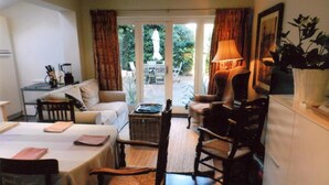 A cozy living room with a sofa, a dining table, and a chair. There is a lamp, a plant, and a window with curtains.