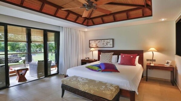 A spacious bedroom with a large bed, two bedside tables, a ceiling fan, and a view of a garden through a sliding glass door.