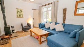 A cozy living room with a blue sofa, a wooden coffee table, a fireplace, and a window with curtains. - Granary - 17th century converted cottage (Looe)