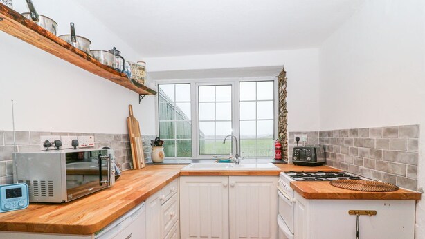 A compact kitchen with white cabinets, a wooden countertop, a built-in oven, a microwave, and a window with a view of greenery. - Granary - 17th century converted cottage (Looe)