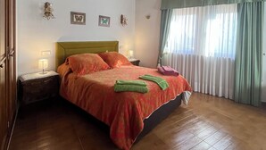 Et soveværelse med træloft, en seng med grønt og rødt sengetæppe, et natbord med lampe og et vindue med gardiner.