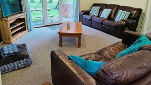A living room with a brown leather sofa, a wooden coffee table, a TV on a wooden stand, and a view through a glass door to a garden. - Little Croft - Spacious cottage in Woodland setting close to fantastic beaches (Truro)