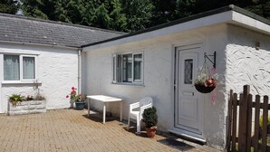 A small, white cottage with a tiled roof, a white door, and a window. There is a white plastic chair and a potted plant near the door. - Little Croft - Spacious cottage in Woodland setting close to fantastic beaches (Truro)