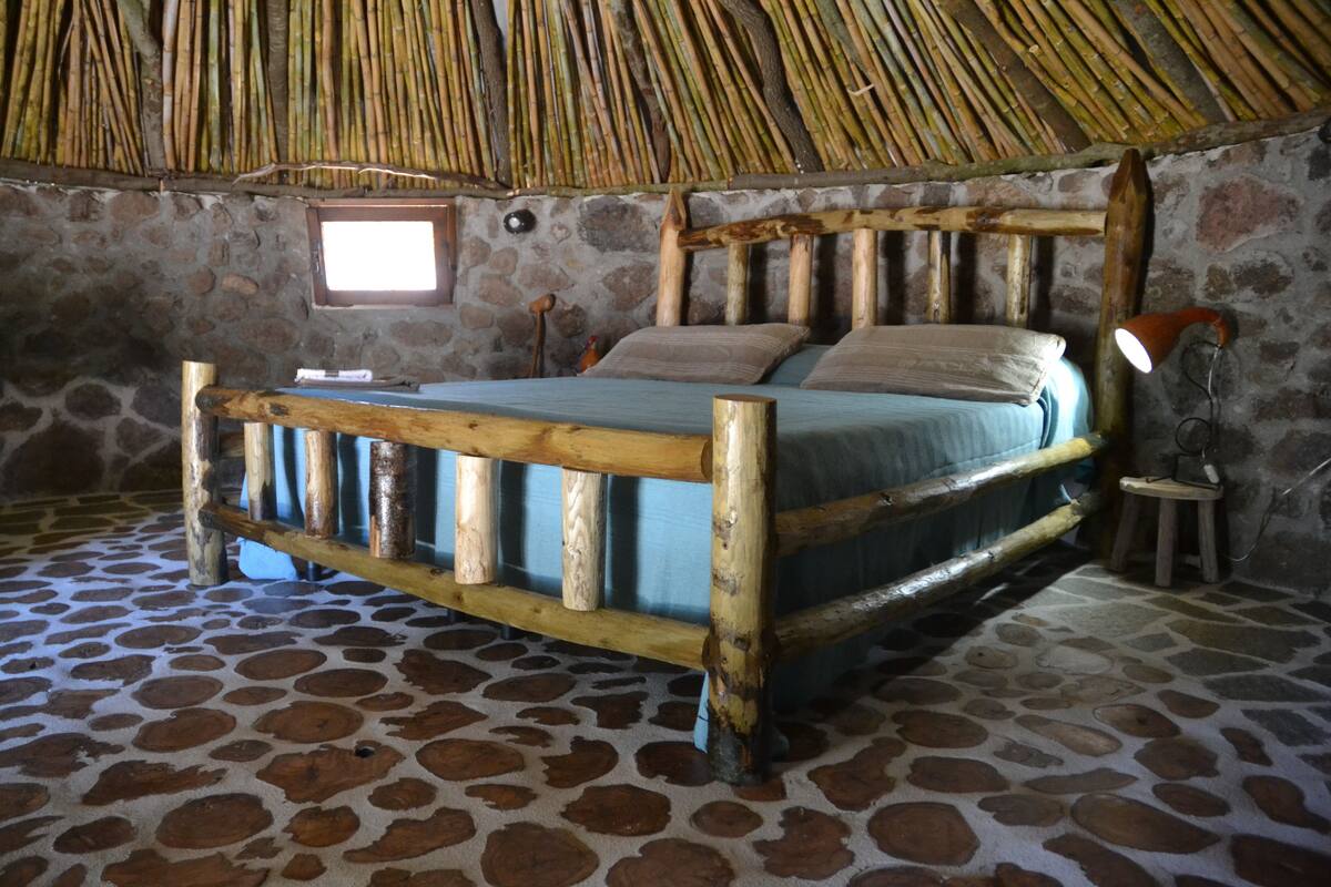 Een rustieke kamer met een stenen vloer, een bed met houten frame, een rieten dak en een klein raam.
