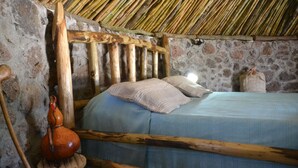 Une pièce rustique avec un mur en pierre, un lit en bois avec une literie bleue et un tabouret en bois surmonté d’une cruche en céramique.