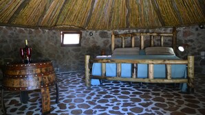 Une pièce avec un toit de chaume, un lit avec un cadre en bois, un mur en pierre, une petite fenêtre et un tonneau en bois.