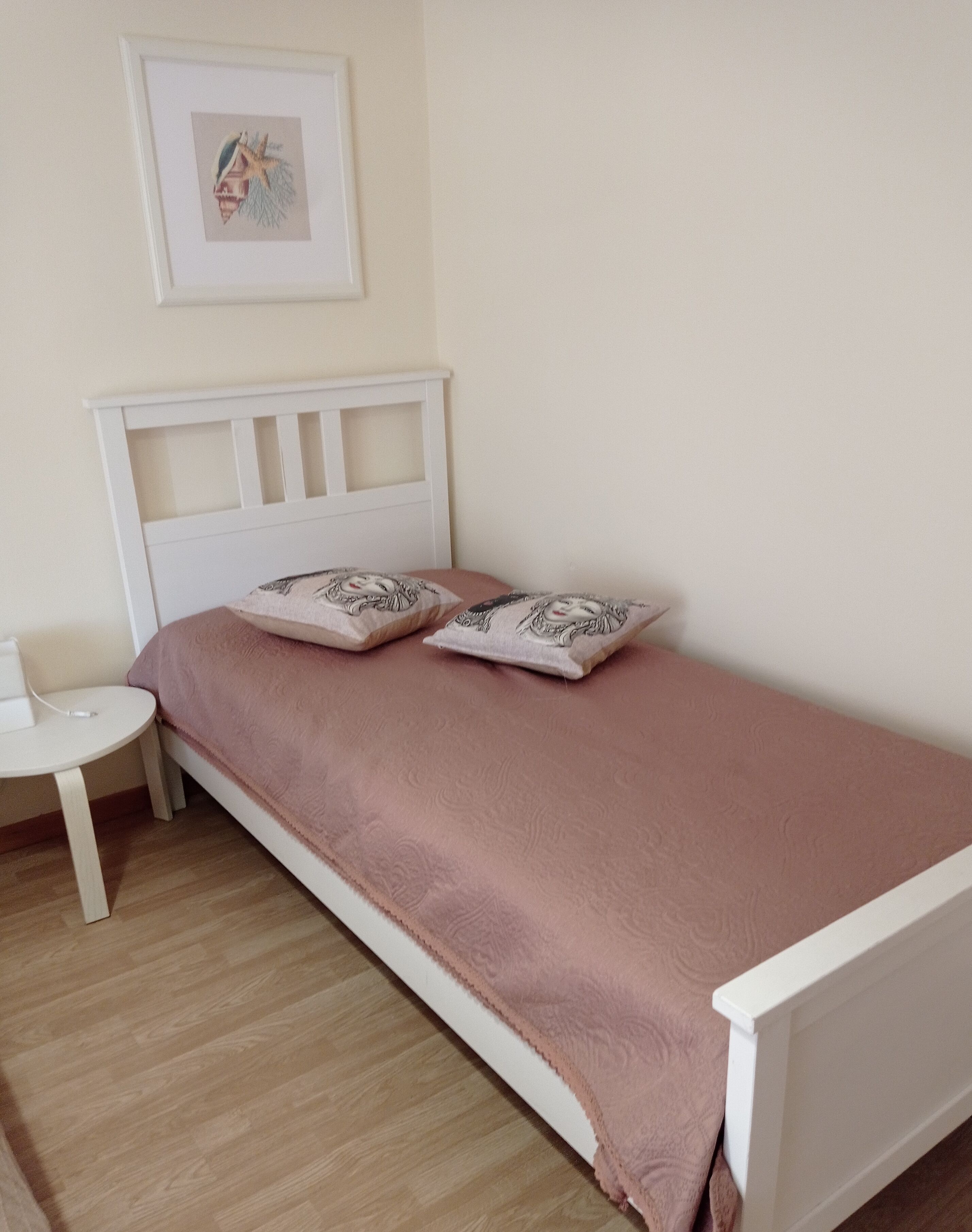 Un lit simple avec une tête de lit blanche et une couvre-lit rose, agrémenté de deux coussins décoratifs. Une petite table de chevet blanche est placée à gauche du lit.