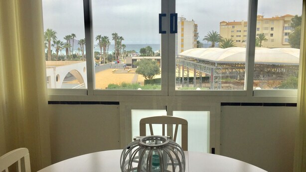 Une pièce dotée d’une grande fenêtre donnant sur une plage et des immeubles. On y trouve une table sur laquelle est posé un objet décoratif en verre.