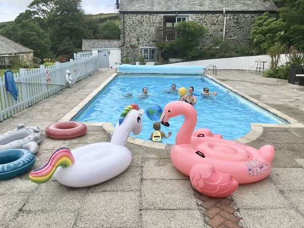 A swimming pool with inflatable swan floats, a stone building, and a white fence in the background. - Self Catering Rural Farm Cottage + Heated Pool Cornwall (Launceston)