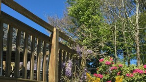 A wooden deck with a stone wall, surrounded by purple wisteria and pink and yellow flowering bushes.