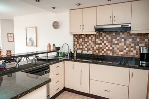 A modern kitchen with beige cabinets, a black countertop, a tiled backsplash, and a stainless steel range hood.