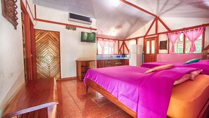 A bedroom with a bed, a wooden desk, a television, and a kitchen area.