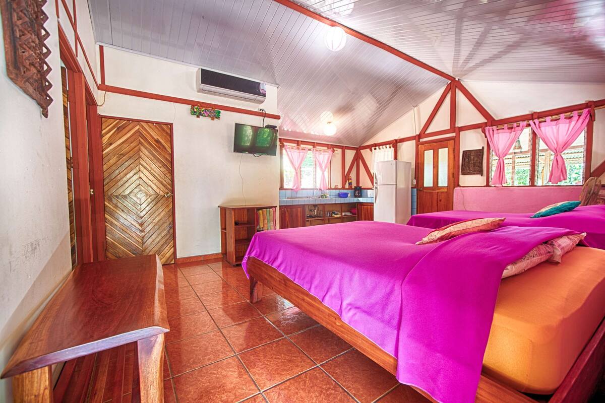 A bedroom with a bed, a wooden desk, a television, and a kitchen area.