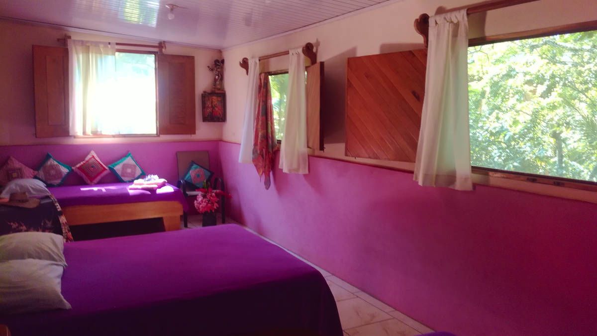A room with two beds, a window with curtains, and a view of greenery outside.