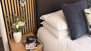 A neatly made bed with white and dark blue pillows, a bedside table with a book and a vase of flowers, and a wall with vertical wooden slats. - Two bed Appt. 10 minutes walk to Lynton . Riverside location. EV charge point (Lynton)