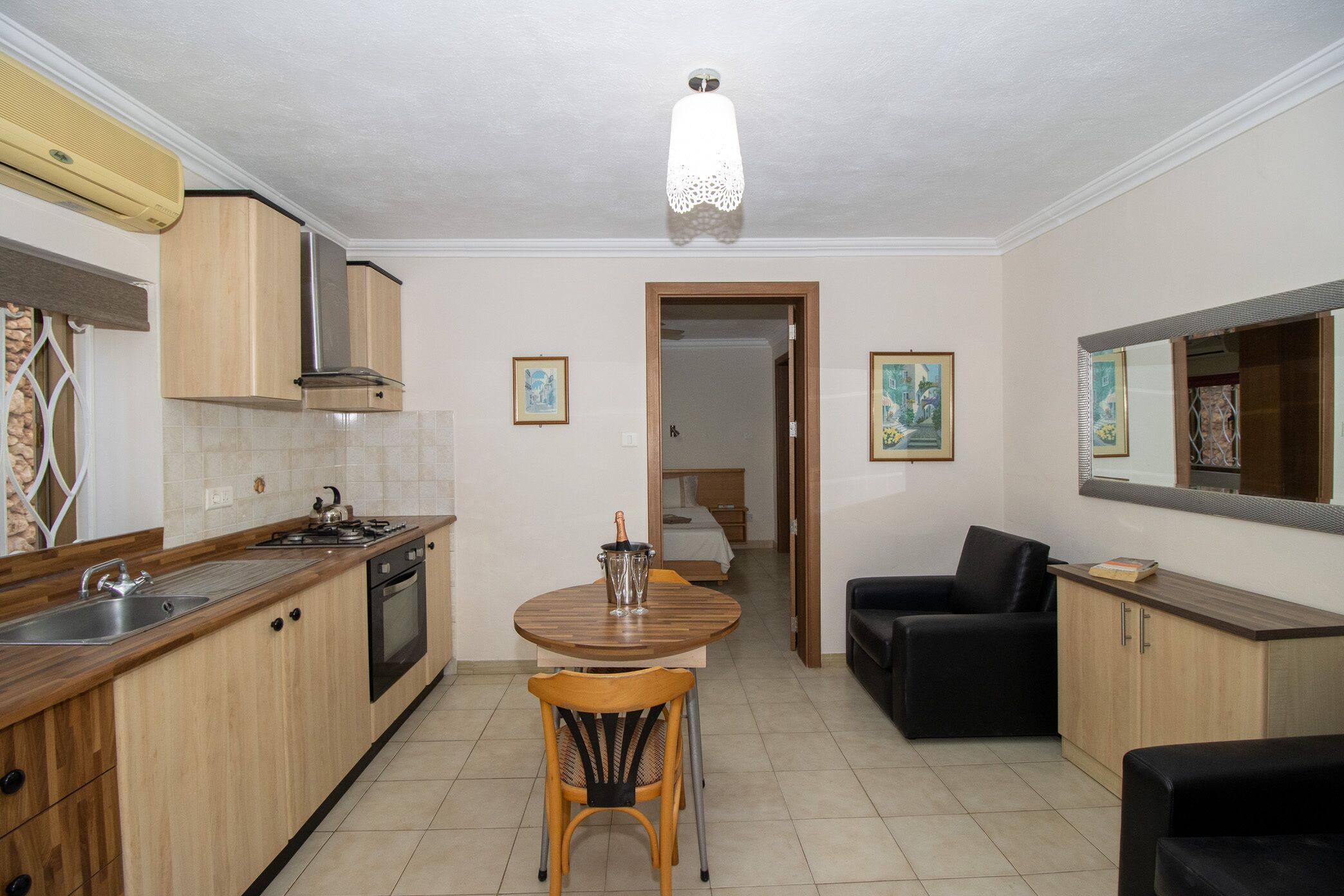 Una cocina compacta con armarios de madera, un lavabo y una estufa. Hay una mesa de comedor con una silla en el centro, y un sofá negro a la derecha.