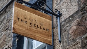 Uma placa de madeira com o texto 'THE CELLAR' pendurada em um suporte de metal numa parede de pedra.