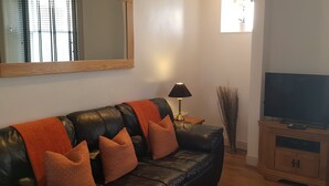 Uma sala de estar com sofá de couro preto, almofadas laranjas, uma TV em um suporte de madeira e uma luminária.