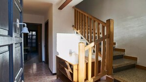 Eine Holztreppe mit Handlauf führt zu einer Aussichtsplattform mit einem weißen Kissen. Eine Tür mit blauem Rahmen und eine weiße Leuchte sind links erkennbar.