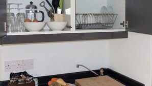 Une cuisine avec un plan de travail noir, un évier et une planche à découper. On y trouve des bols, un panier et un égouttoir sur les étagères.