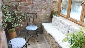 Un petit patio clos, avec un mur en briques, une table en verre, deux chaises, un banc rembourré et une vue sur un canal, visible à travers une fenêtre.