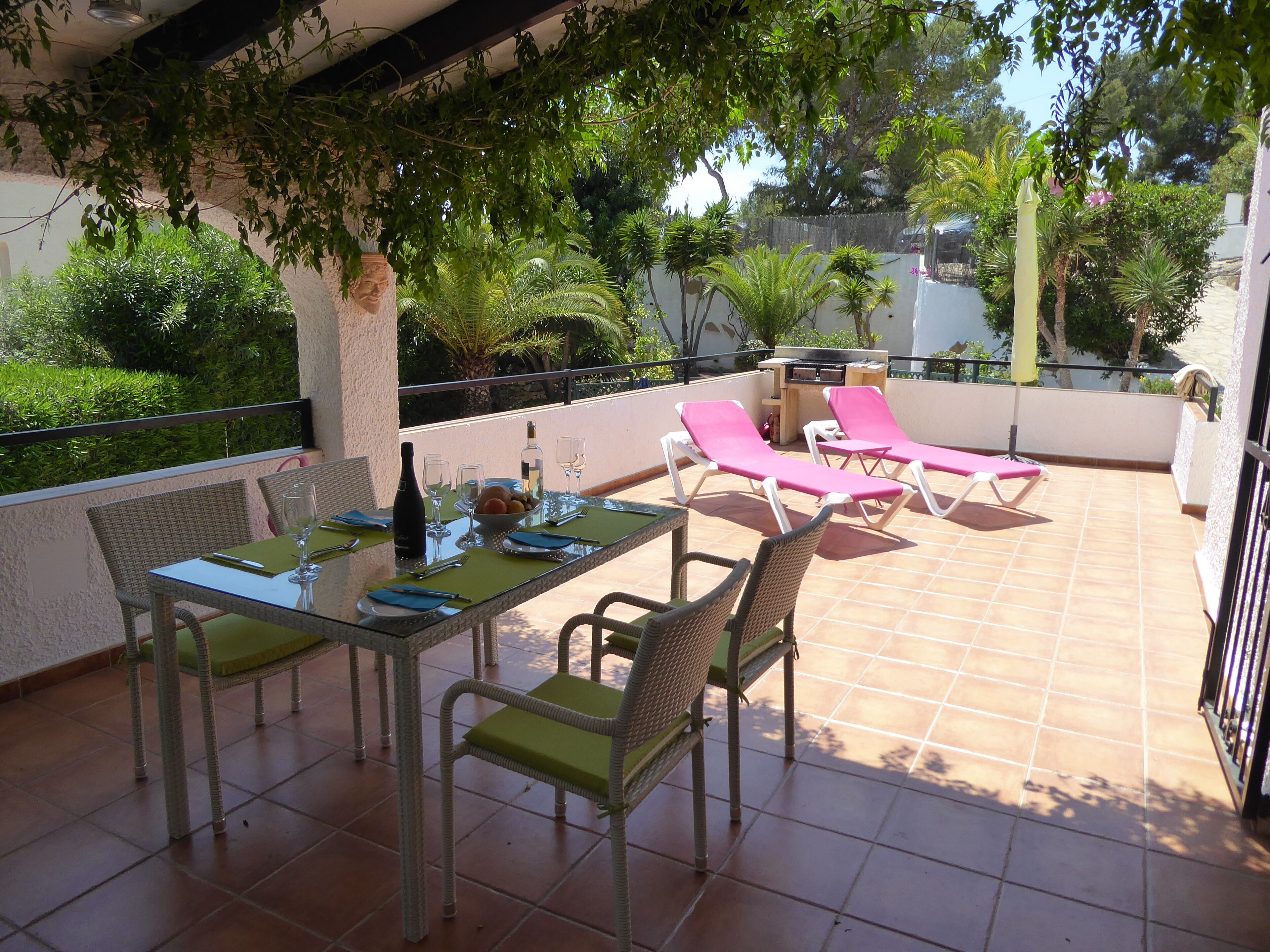 Une terrasse extérieure avec une table à manger pour deux personnes, entourée de chaises en osier et d’un coin salon avec deux transats roses.