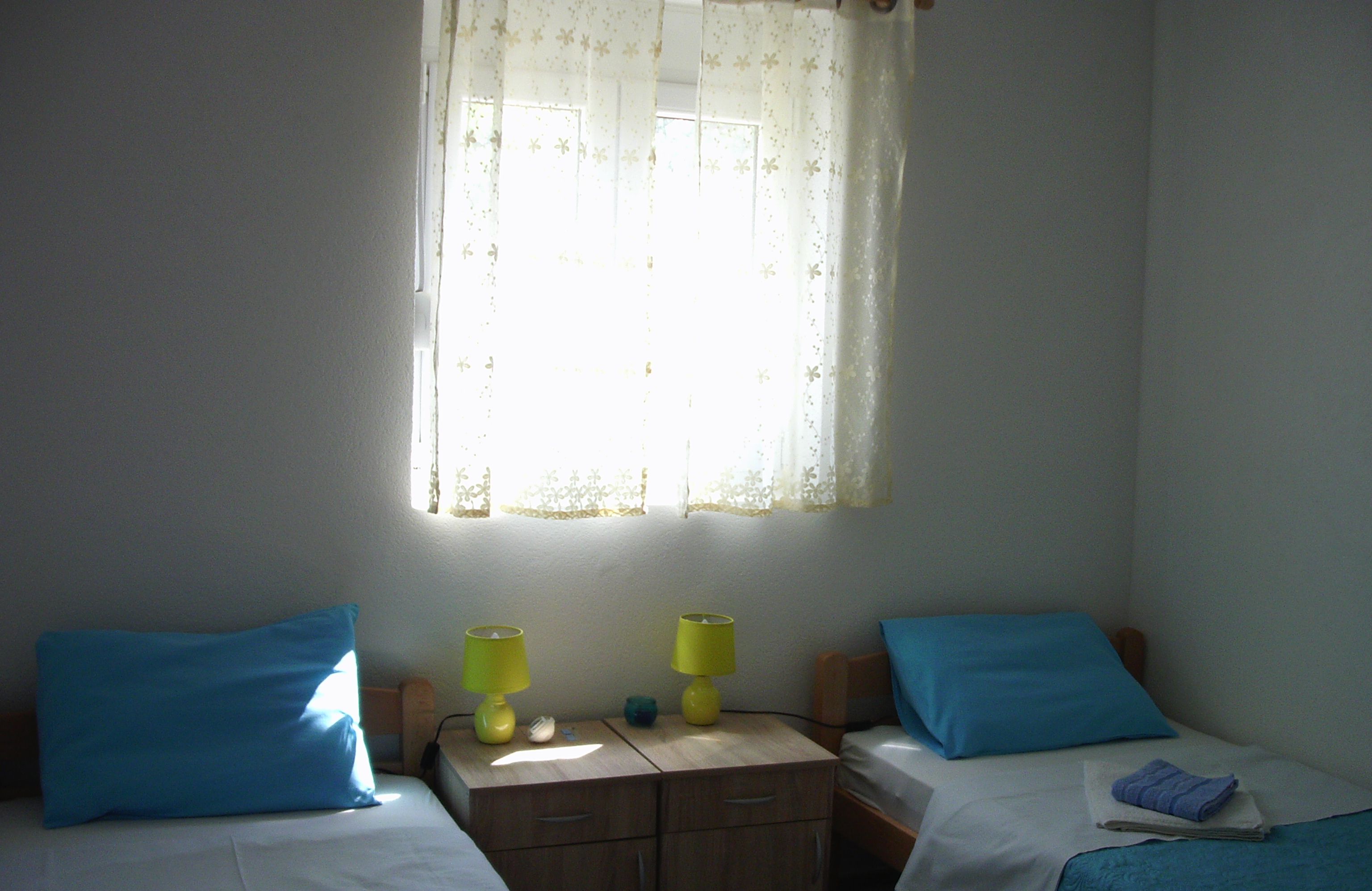 Una habitación con dos camas, cada una con cubrecama y almohada azul. Entre las camas, un mesita de noche de madera con dos lámparas amarillas. Se ve una ventana con cortina estampada de flores, y se muestra una marca de fecha y hora.