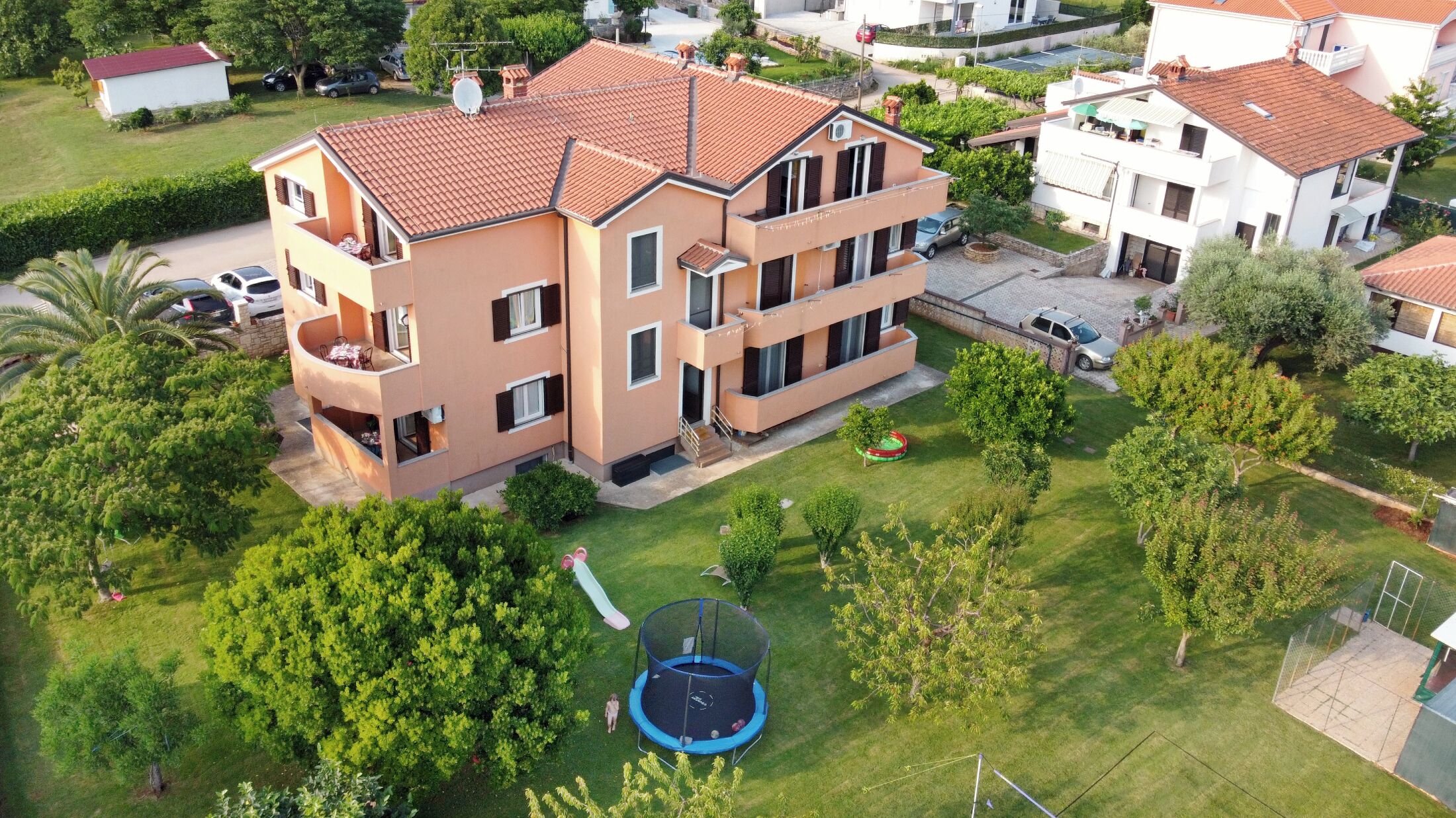 A multi-story residential building with a tiled roof, a visible balcony, and a well-maintained lawn with a trampoline.