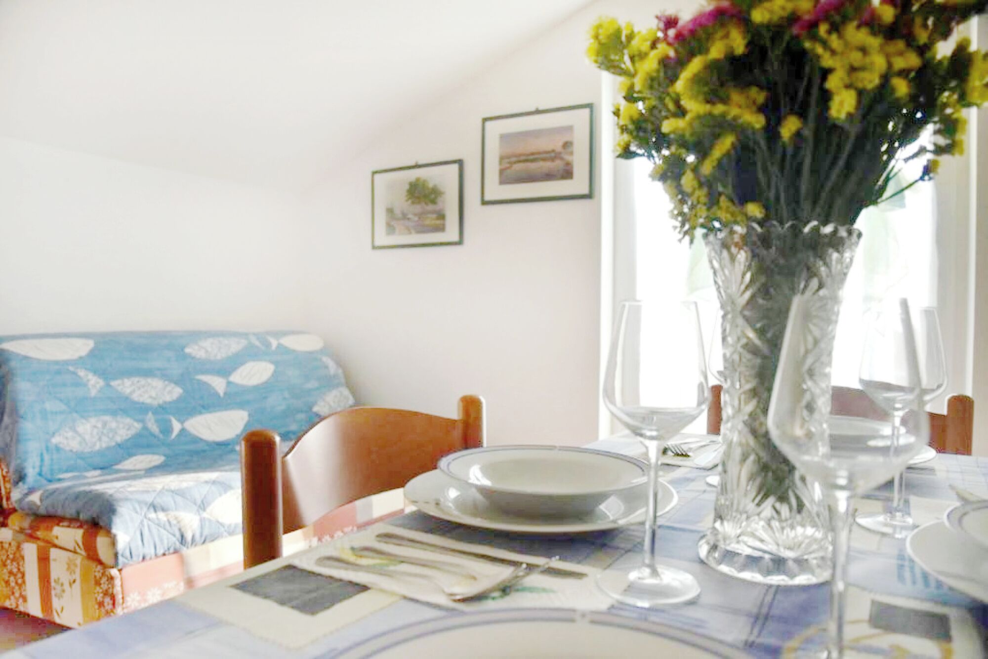 Una tavola imbandita con piatti, posate e un vaso di fiori. Su una sedia c'è un cuscino blu decorato e al muro una fotografia incorniciata.