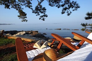 A beachside lounge with wooden chairs and straw hats, overlooking a calm sea and rocky shoreline.